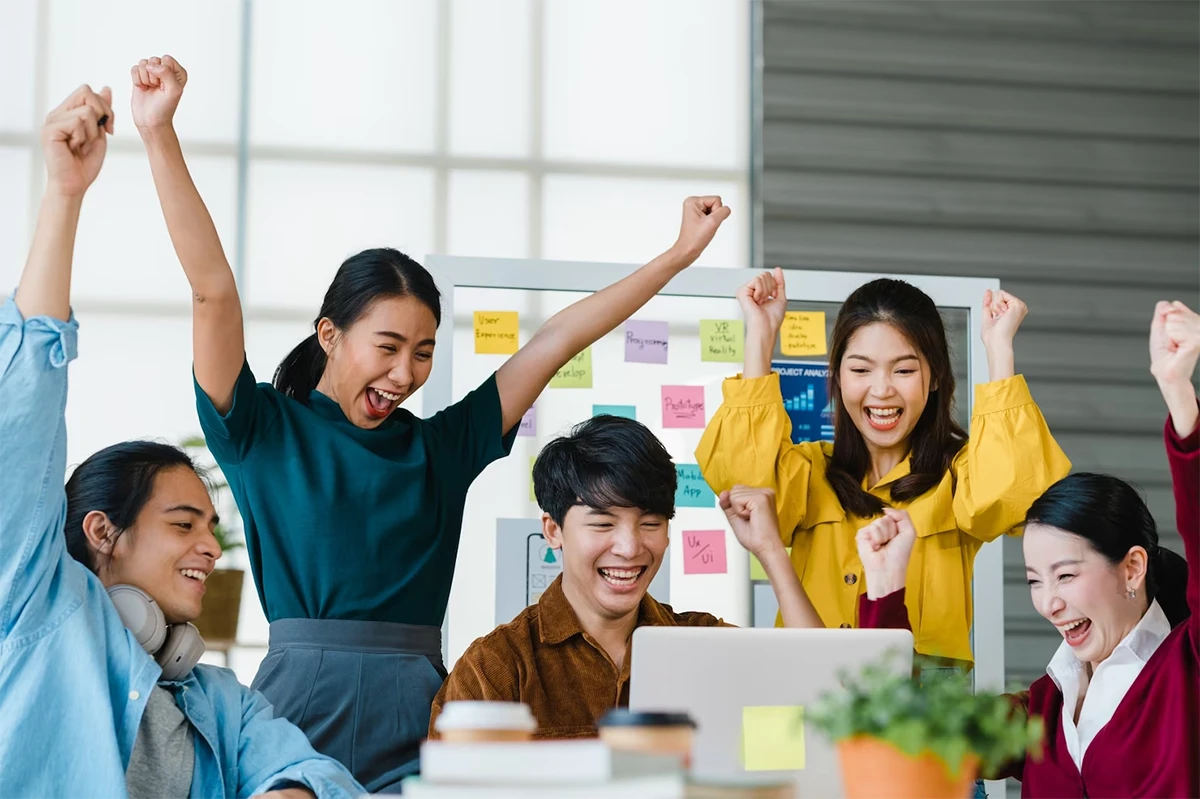 Kenapa Harus Belajar Bahasa Inggris? Ini Manfaatnya untuk Kehidupan Kamu 1 Lima orang dewasa muda bersorak dan mengangkat tangan mereka dengan penuh kegembiraan di sekitar sebuah laptop di sebuah kantor modern. Mereka duduk dan berdiri di dekat papan tulis putih yang dipenuhi catatan tempel berwarna-warni, merayakan bersama-sama. Cangkir kopi dan buku catatan terlihat di atas meja.